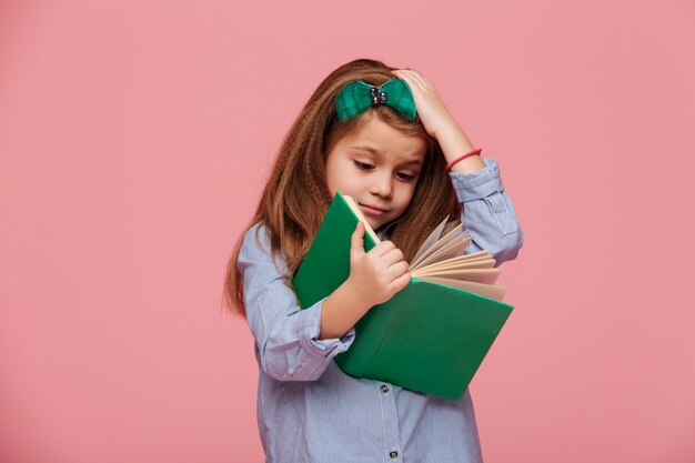 Imagen de niña divertida en camisa agarrando su cabeza mientras leía el libro cansada de estudiar