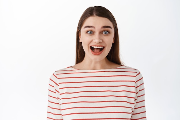 Imagen de una niña emocionada y sorprendida que escucha una buena noticia increíble, se queda boquiabierto y se queda asombrado, impresionado por la cámara, fascinado por algo increíble, de pie sobre un fondo blanco.