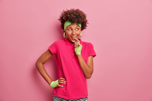 Foto gratuita la imagen de la niña de pelo rizado tiene una expresión alegre y astuta, se ve con la intención de implementar el plan, vestida con ropa informal