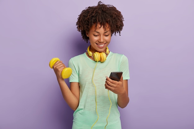 Imagen de niña satisfecha en forma rizada de pelo oscuro elige la pista en la lista de reproducción, escucha música a través de auriculares, levanta el brazo con mancuernas, tiene entrenamiento activo, aislado en la pared violeta. Concepto de culturismo