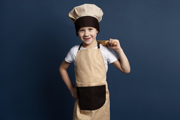 Imagen de un niño de 7 años de ojos azules alegre cocinero con uniforme de chef sosteniendo un rodillo en su hombro, alegrándose mientras amasa la masa para galletas de jengibre, mirando a la cámara con una sonrisa feliz