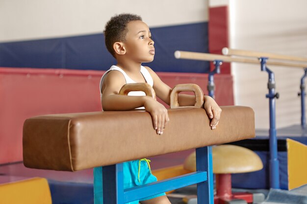 Imagen de un niño afroamericano de diez años en camiseta blanca de pie junto a un caballo con arcos en el gimnasio