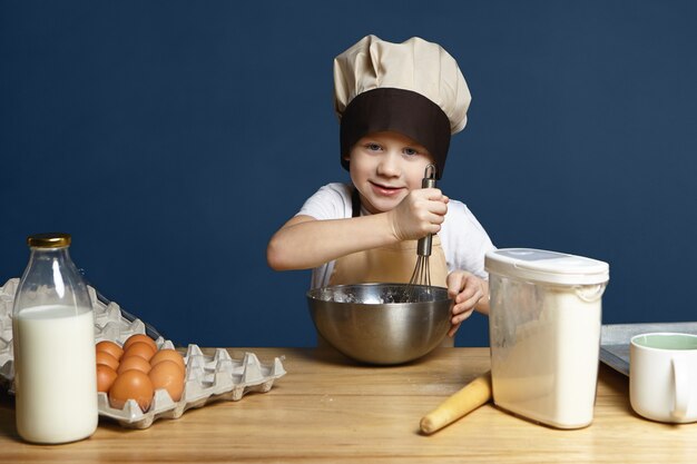 Imagen de niño con delantal y gorra de chef batiendo ingredientes en un tazón de metal mientras cocina panqueques, galletas u otros pasteles, de pie en la mesa de la cocina con huevos, leche, harina y rodillo