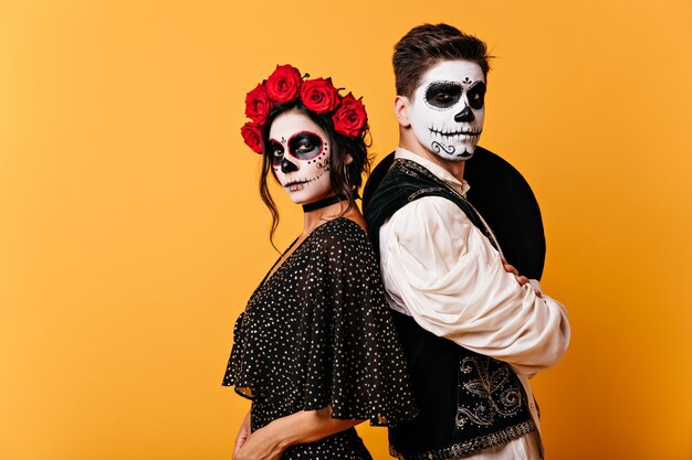 Imagen de una orgullosa pareja mexicana en ropas tradicionales con caras pintadas. Chica con rosas en el pelo posa con joven con sombrero a la espalda.