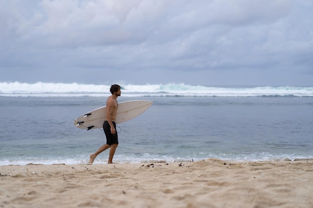 Imagen paisajística de un surfista masculino ocupado caminando por la playa al amanecer mientras lleva su tabla de surf bajo el brazo con las olas rompiendo en el fondo. Joven surfista masculino guapo en el océano