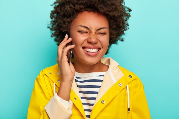 Imagen recortada de atractiva mujer de piel oscura con cabello afro, se ríe de la divertida historia de amigos, sostiene el teléfono celular, vestida con un impermeable amarillo