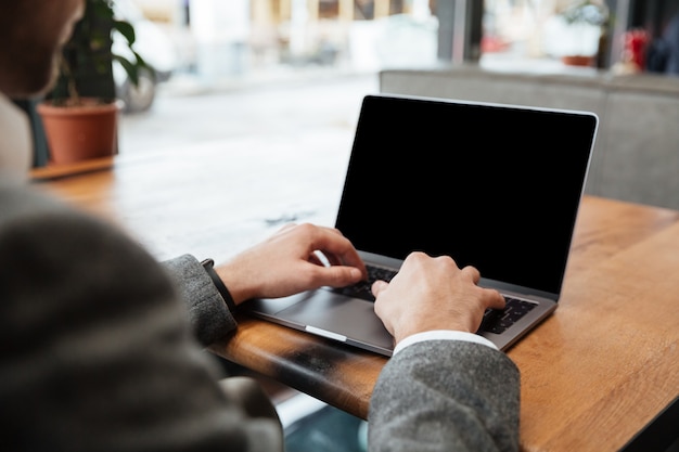 Imagen recortada del empresario sentado junto a la mesa en la cafetería y escribiendo en la computadora portátil