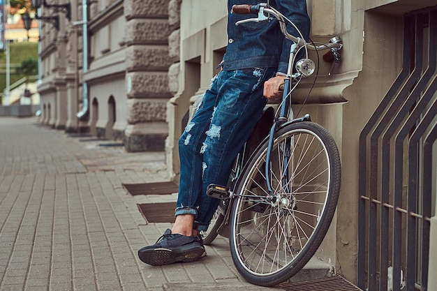 Imagen recortada de un hombre de moda con ropa elegante apoyado contra una pared con bicicleta de ciudad en una calle.