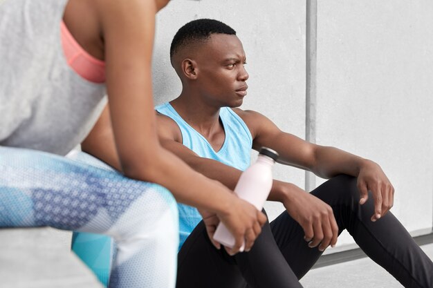 Imagen recortada de un hombre de piel oscura que se sienta cansado, su pareja posa cerca con una botella de agua fría, hacen deporte juntos, llevan un estilo de vida activo, tienen entrenamientos regulares en el gimnasio o al aire libre, usan ropa deportiva