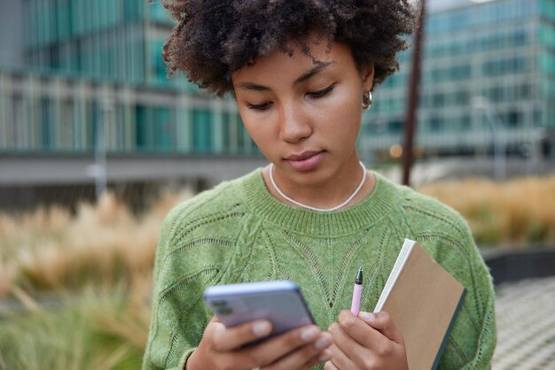 Imagen recortada de un modelo femenino serio concentrado en la pantalla del teléfono inteligente que lee información en línea sostiene un diario y un bolígrafo para escribir notas usa un jersey casual que se encuentra solo al aire libre