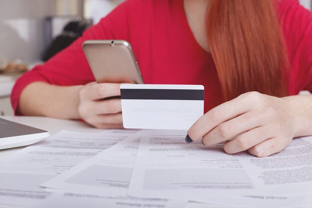 La imagen recortada de una mujer de jengibre irreconocible trabaja con documentación, intenta relajarse y hacer compras en línea, usa un teléfono inteligente y una tarjeta de plástico, paga por la compra. Personas, pagos, comercio electrónico.