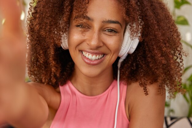 Imagen recortada de una mujer negra de raza mixta alegre con cabello nítido, hace un retrato selfie, escucha la pista de audio en los auriculares, está de buen humor, tiene tiempo libre después de las clases, disfruta de un audiolibro emocionante