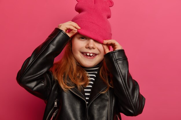 La imagen recortada de la niña se ve desde el sombrero, esconde la cara, viste una elegante chaqueta de cuero negro, vestida con ropa de moda tiene un aspecto positivo y ambicioso aislado en una pared rosa. Niños, emociones, estilo