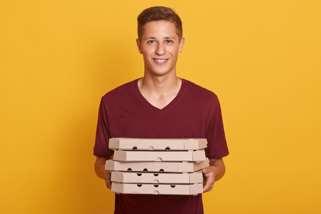 Imagen del repartidor hermoso alegre que lleva la camiseta casual de Borgoña, sosteniendo la pila de cajas de pizza en las manos y mirando directamente a la cámara aislada en el estudio amarillo. Concepto de comida chatarra.