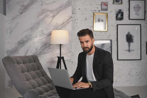 Imagen de serio guapo experimentado joven profesional masculino caucásico que trabaja en su elegante y moderno apartamento, sentado en el sofá con ropa formal, mecanografía usando una computadora portátil abierta