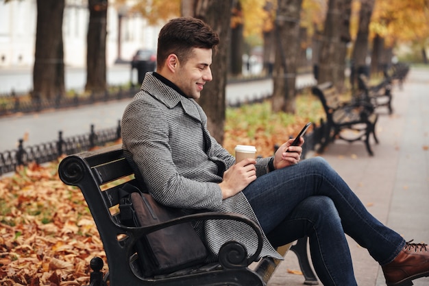 Imagen de un sonriente hombre moreno con abrigo y jeans tomando café para llevar y usando su teléfono móvil, mientras está sentado en el banco en el parque