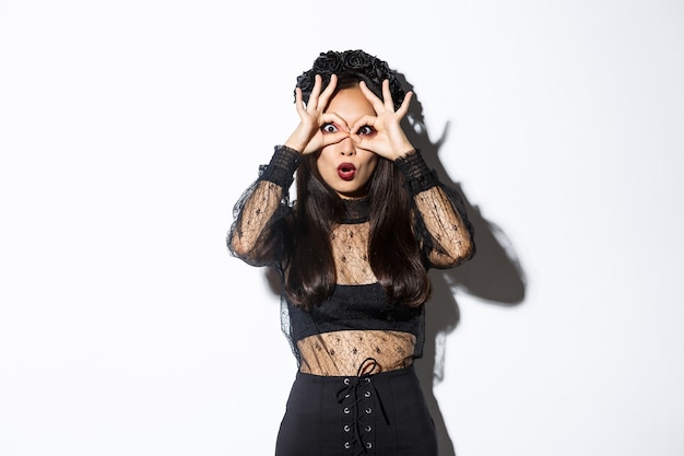 Foto gratuita imagen de sorprendida linda mujer asiática vestida con traje de bruja, haciendo gafas con los dedos sobre los ojos y mirando a través de ellos impresionado