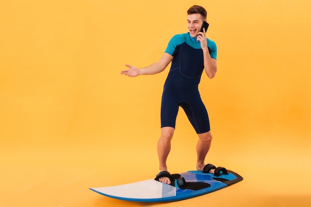 Imagen de surfista feliz sorprendido en traje de neopreno con tabla de surf y hablando por teléfono inteligente