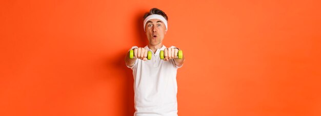 Imagen de un tipo activo de fitness de mediana edad haciendo ejercicios deportivos con pesas y sonriendo emocionado de pie sobre un fondo naranja