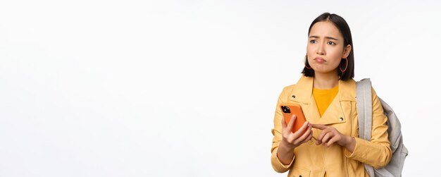 Imagen de una turista viajera feliz con mochila mirando el teléfono inteligente usando la aplicación de ruta de teléfono móvil de pie sobre fondo blanco Copiar espacio