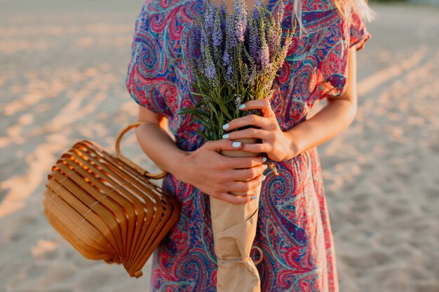 Imagen de verano al aire libre de hermosa mujer rubia romántica en vestido colorido caminando por la playa con ramo de lavanda.