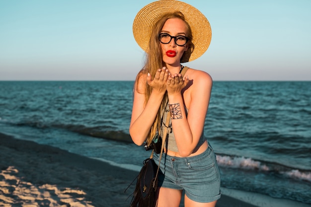 Imagen de verano al aire libre de hermosa mujer rubia con sombrero de paja caminando cerca del mar.