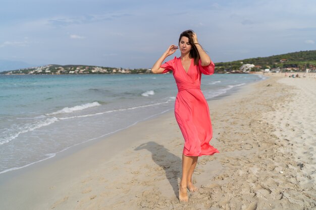 Imagen de verano de feliz mujer sexy en vestido rosa precioso posando en la playa. De longitud completa.