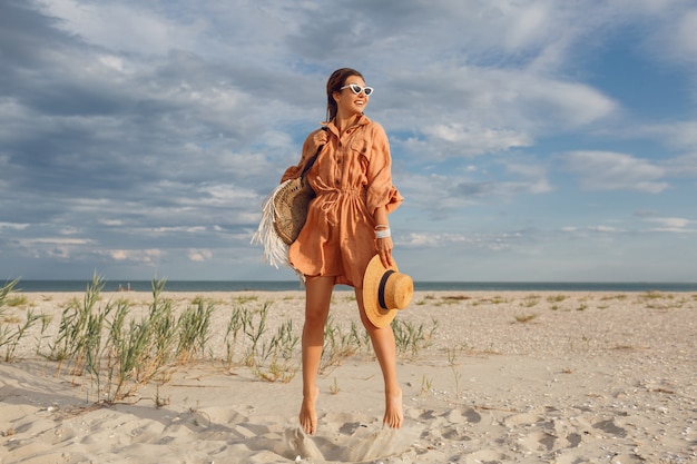 Imagen de verano de hermosa mujer morena en vestido de lino de moda saltando y jugando, sosteniendo una bolsa de paja. Chica muy delgada disfrutando de los fines de semana cerca del océano. Longitud total.