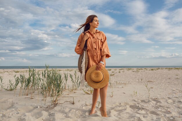 Imagen de verano de hermosa mujer morena en vestido de lino de moda saltando y jugando, sosteniendo una bolsa de paja. Chica muy delgada disfrutando de los fines de semana cerca del océano.