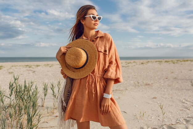 Imagen de verano de hermosa mujer morena en vestido de lino de moda, sosteniendo una bolsa de paja. Chica muy delgada disfrutando de los fines de semana cerca del océano.