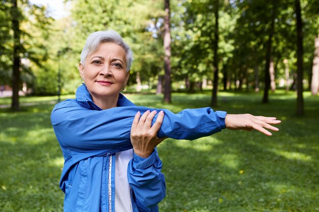 Imagen de verano de mujer jubilada activa sana sonriendo, estirando los músculos del brazo después de correr el entrenamiento al aire libre, posando en el bosque. Concepto de salud, bienestar, edad, personas, deportes y actividad.