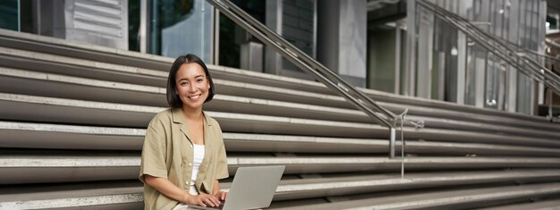 Imagen vertical de una chica asiática sentada con una computadora portátil tomando café en las escaleras de la universidad joven estudiante