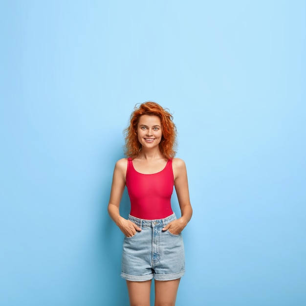 Foto gratuita imagen vertical de delgada joven jengibre vestida con chaleco rojo y pantalones cortos de mezclilla, mantiene las manos en los bolsillos