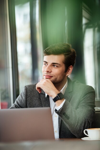 Imagen vertical del empresario pensativo sentado junto a la mesa en la cafetería con computadora portátil y apartar la mirada