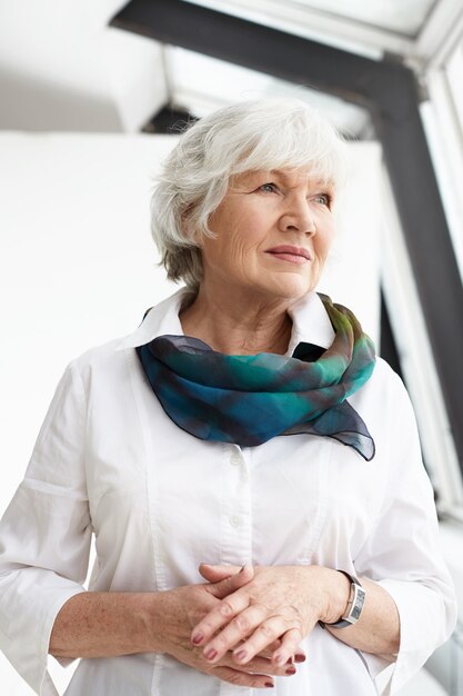 Imagen vertical de la encantadora abuela europea elegante con ropa elegante y elegante y accesorios que pasan tiempo en el interior, pensando en algo, mirando a otro lado con expresión seria pensativa