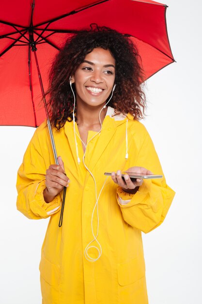 Imagen vertical de feliz mujer africana en gabardina escondiéndose bajo el paraguas y escuchando música en su teléfono mientras mira lejos sobre blanco