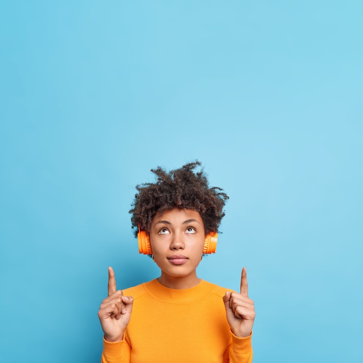 Foto gratuito la imagen vertical de una hermosa mujer afroamericana seria concentrada arriba indica hacia arriba muestra espacio de copia para su contenido publicitario o logotipo escucha música a través de auriculares. coloque su texto aquí