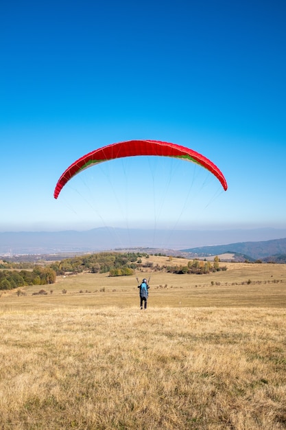 Imagen vertical de un hombre volando con un paracaídas rojo rodeado de vegetación bajo un cielo azul