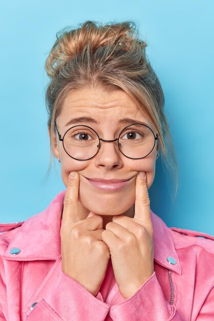 Imagen vertical de una joven y bonita mujer europea que fuerza una sonrisa alegre que mantiene los dedos índices cerca de la comisura de los labios y trata de ser feliz lleva una chaqueta rosa de gafas redondas aislada sobre fondo azul.