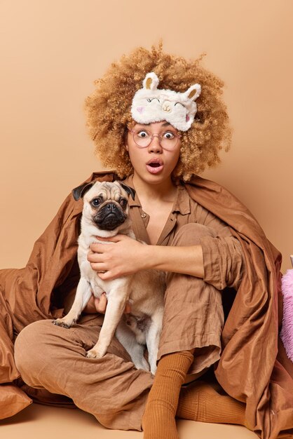 Imagen vertical de una joven de cabello rizado sorprendida que se queda en la cama envuelta en un edredón reacciona ante increíbles juegos de noticias con un perro pug aislado sobre un fondo beige Una modelo femenina asustada se prepara para dormir