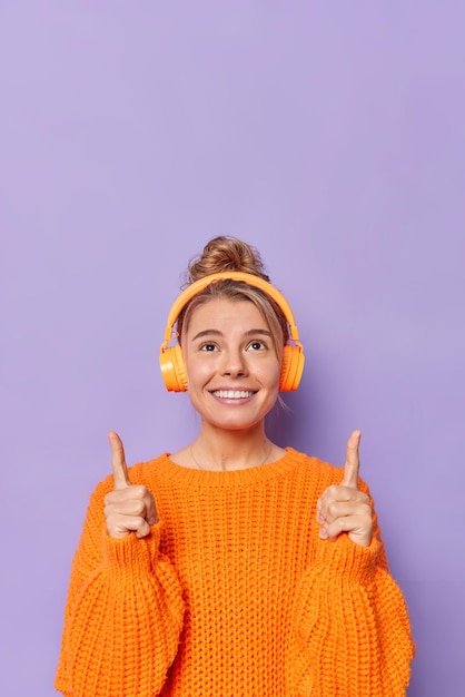 Imagen vertical de una joven feliz y sonriente que señala con el dedo índice por encima de la cabeza y escucha música a través de auriculares vestidos con suéter de punto aislado sobre fondo morado con espacio en blanco para colocar texto.