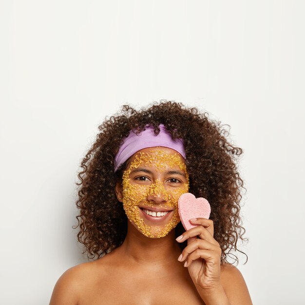 La imagen vertical de una mujer alegre y alegre aplica un exfoliante de sal marina para absorber la suciedad y limpiar los puntos oscuros en la cara, mantiene una buena hidratación, sostiene una esponja en forma de corazón cerca de la mejilla, invierte las células de la piel