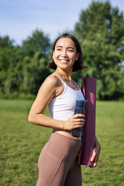 Imagen vertical de una mujer asiática sana y en forma posando en el parque sosteniendo una botella de agua y una estera de goma para yoga