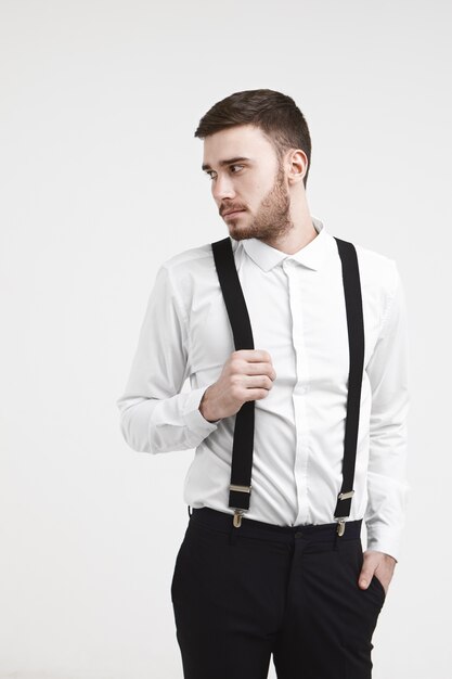 Imagen vertical recortada del apuesto joven empleado masculino caucásico de moda con barba recortada y elegante corte de pelo posando en la pared del estudio, mirando a otro lado, sosteniendo la mano en sus tirantes negros