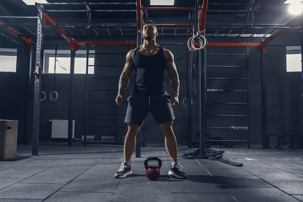 Imparable. Joven atleta caucásico muscular practicando sentadillas en el gimnasio con el peso. Modelo masculino haciendo ejercicios de fuerza, entrenando la parte inferior del cuerpo. Bienestar, estilo de vida saludable, concepto de culturismo.