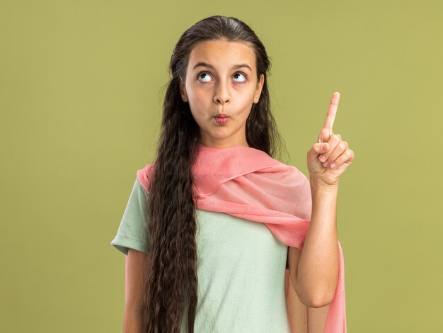 Impresionada adolescente vistiendo un chal mirando y apuntando hacia arriba con los labios fruncidos aislados en la pared verde oliva