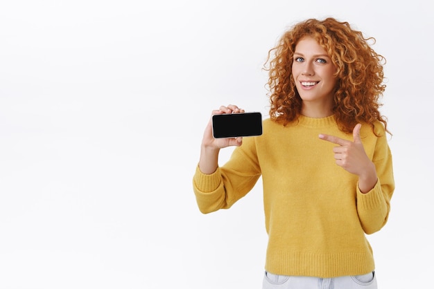 Foto gratuita impresionada, alegre y atractiva mujer pelirroja rizada con suéter amarillo sosteniendo un teléfono inteligente apuntando horizontalmente a la pantalla móvil y sonriendo invitándote a probar esta aplicación de recomendación de sorteo de oferta de juego