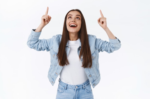 Foto gratuita impresionada y alegre chica morena que mira asombrada y asombrada, contempla algo hermoso y fresco, levanta la cabeza apuntando hacia el anuncio superior, sonriendo divertida, de pie fondo blanco