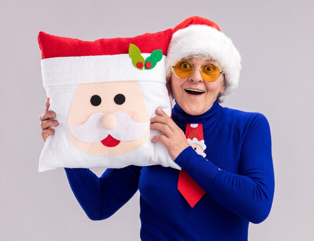 Impresionada anciana en gafas de sol con gorro de Papá Noel y corbata de santa sosteniendo almohada de santa aislado en la pared blanca con espacio de copia