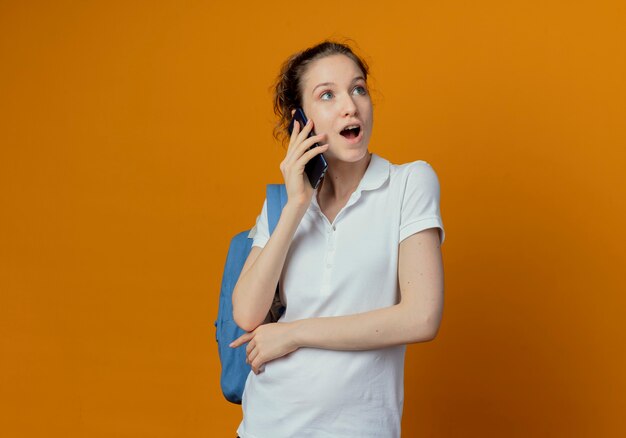 Impresionada bastante joven estudiante vistiendo bolsa trasera mirando al lado hablando por teléfono aislado sobre fondo naranja